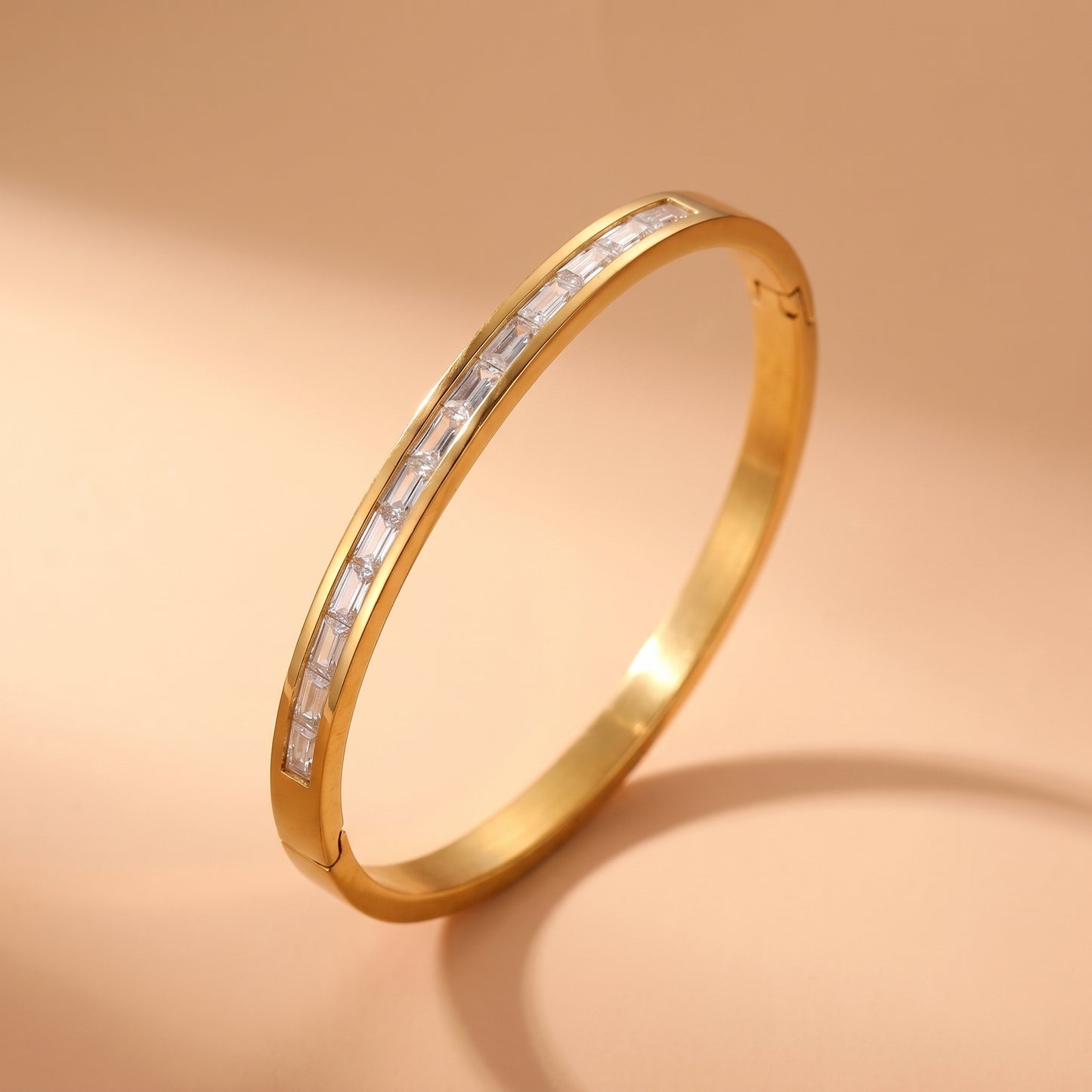 High quality gold Aria Bracelet featuring channel-set clear baguette crystals resting on travertine stone, highlighting its tarnish-free and skin friendly finish, displayed on a warm beige background.