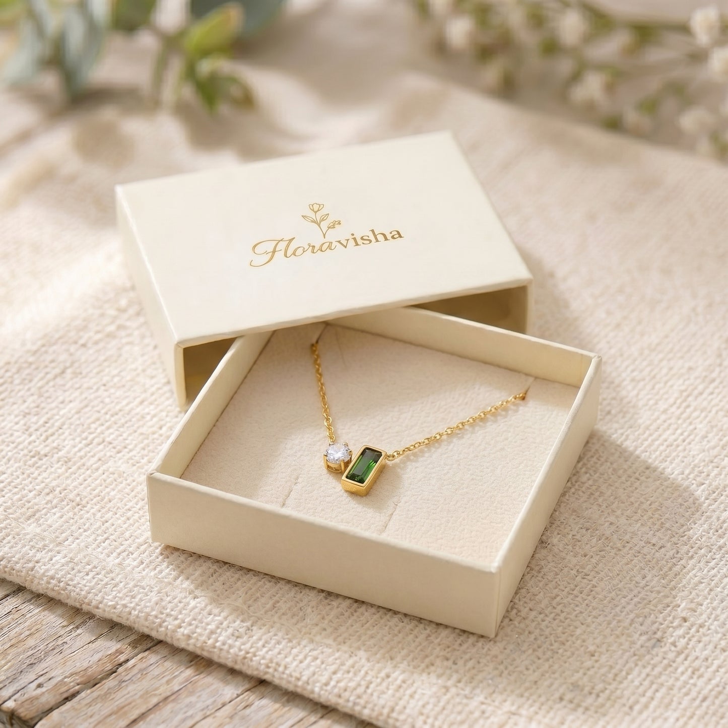 Gold necklace with a green pendant in an open jewelry box labeled 'Floravisha' on a textured surface.