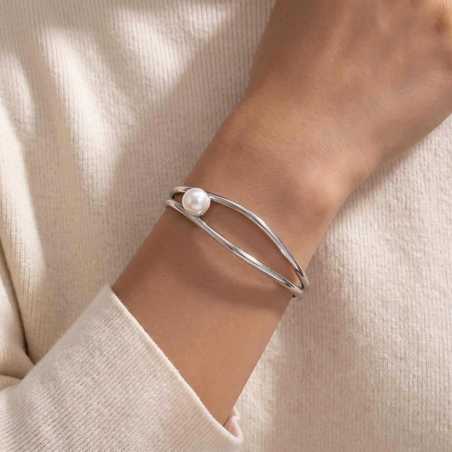 A close-up of a person's wrist wearing a silver double-band cuff bracelet with a single white pearl accent, set against a soft cream-colored knit top.
