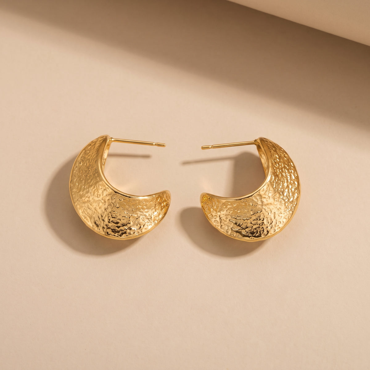 Tarnish-free Aurelia gold tapered crescent half-hoop earrings displaying hammered molten textures and stud posts on a smooth beige background.