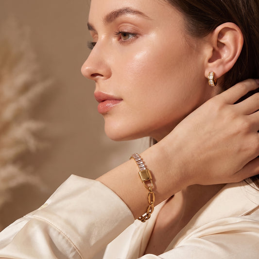 Side profile of a woman wearing the skin friendly Elysée gold paperclip lock bracelet paired with matching gold baguette hoops and a cream silk blouse.