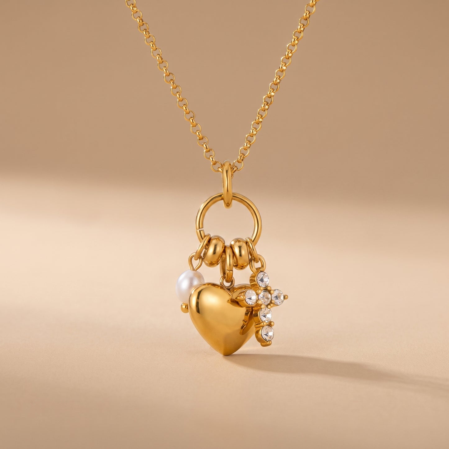 Sleek gold Amara Necklace featuring a gold heart pendant, lustrous pearl drop, and crystal cross charm on a warm beige background, highlighting its tarnish-free finish. 