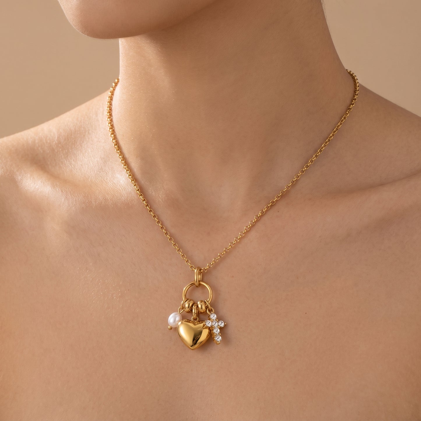 Sleek gold Amara Necklace featuring a gold heart pendant, lustrous pearl drop, and crystal cross charm, highlighting its tarnish-free finish, shown worn on a model's neck against a warm beige background.