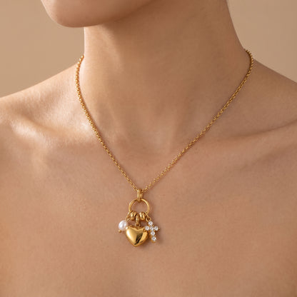 Sleek gold Amara Necklace featuring a gold heart pendant, lustrous pearl drop, and crystal cross charm, highlighting its tarnish-free finish, shown worn on a model's neck against a warm beige background.