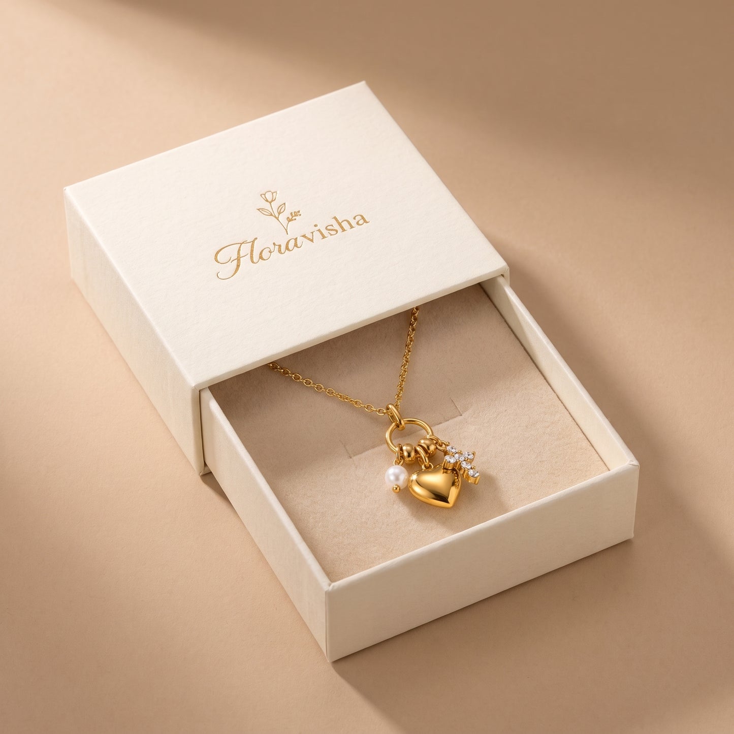 Sleek gold Amara Necklace featuring a gold heart pendant, lustrous pearl drop, and crystal cross charm on a clean solid beige background, highlighting its tarnish-free finish presented in an open cream-colored jewelry box. 