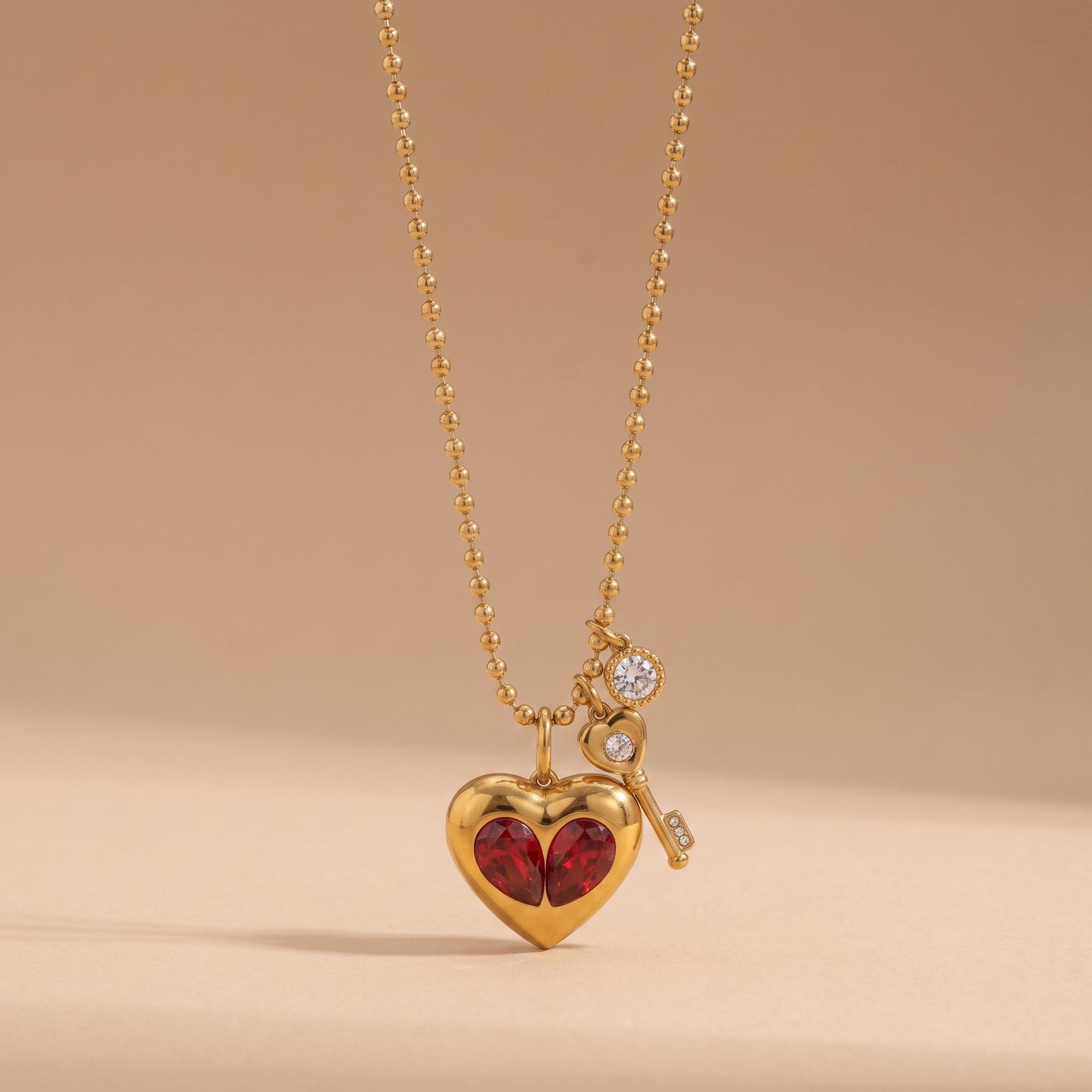 Sleek gold Amour Necklace featuring red crystal heart inserts, a golden key charm, and a clear crystal drop on a warm beige background.