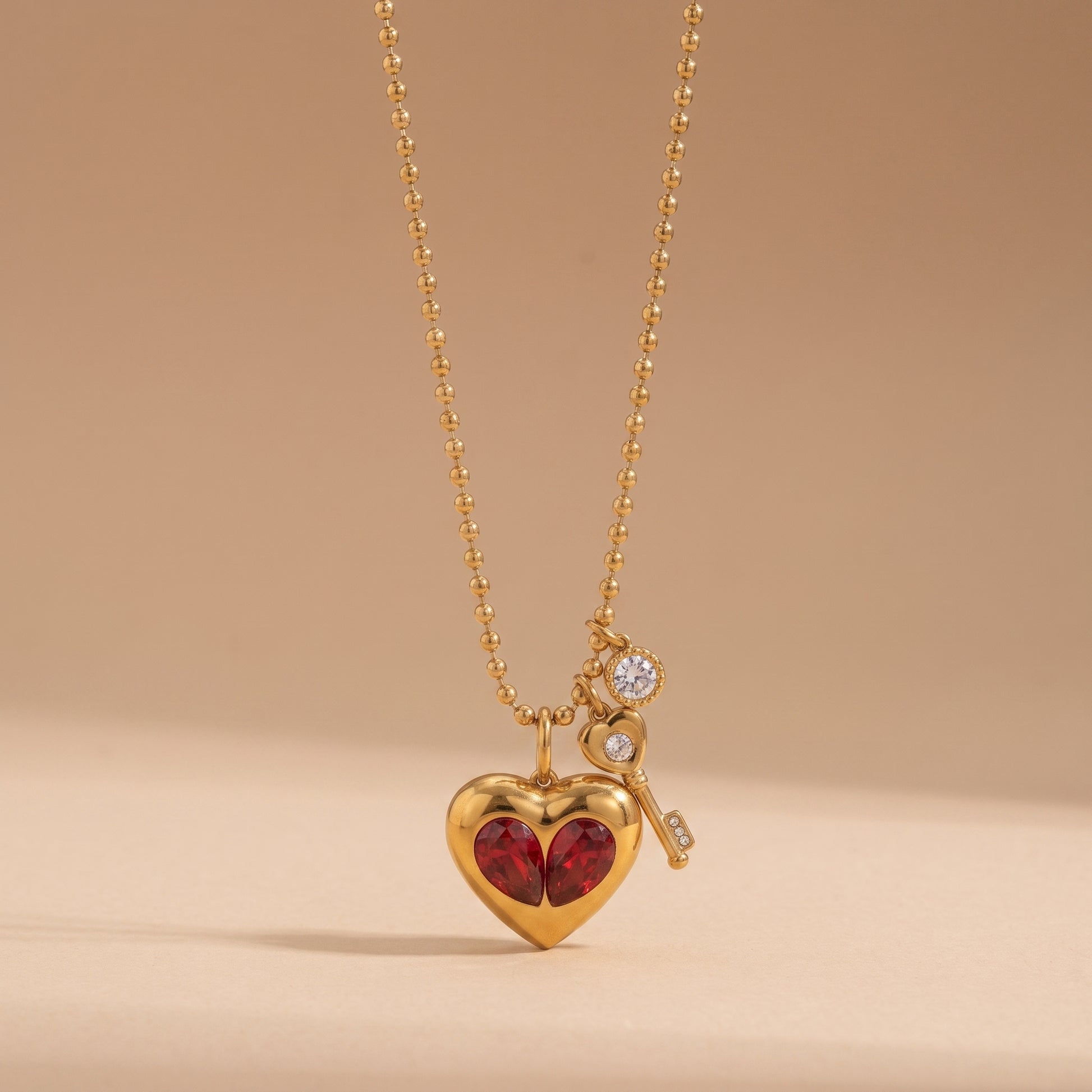 Sleek gold Amour Necklace featuring red crystal heart inserts, a golden key charm, and a clear crystal drop on a warm beige background.
