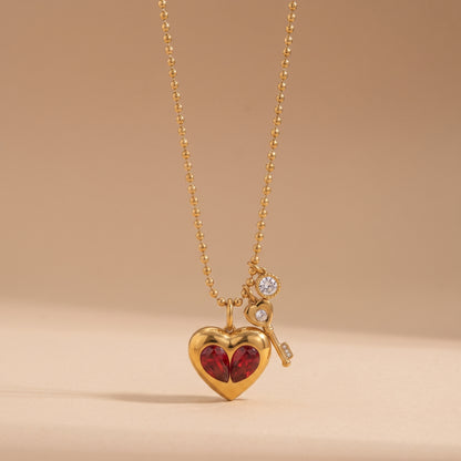 Sleek gold Amour Necklace featuring red crystal heart inserts, a golden key charm, and a clear crystal drop on a warm beige background.