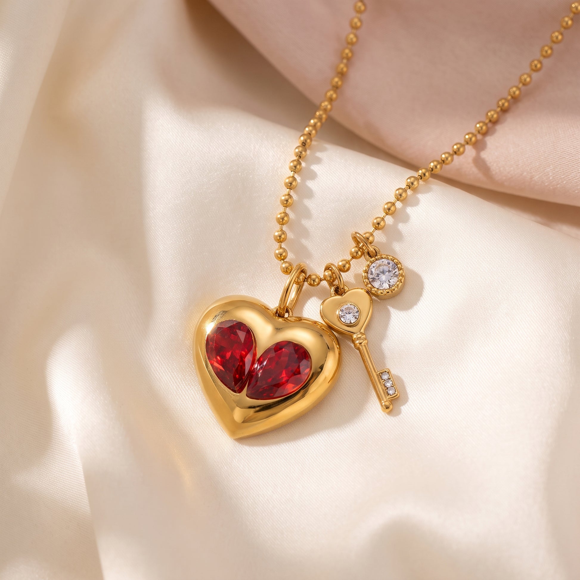 Sleek gold Amour Necklace featuring red crystal heart inserts, a golden key charm, and a clear crystal displayed on draped cream satin fabric.