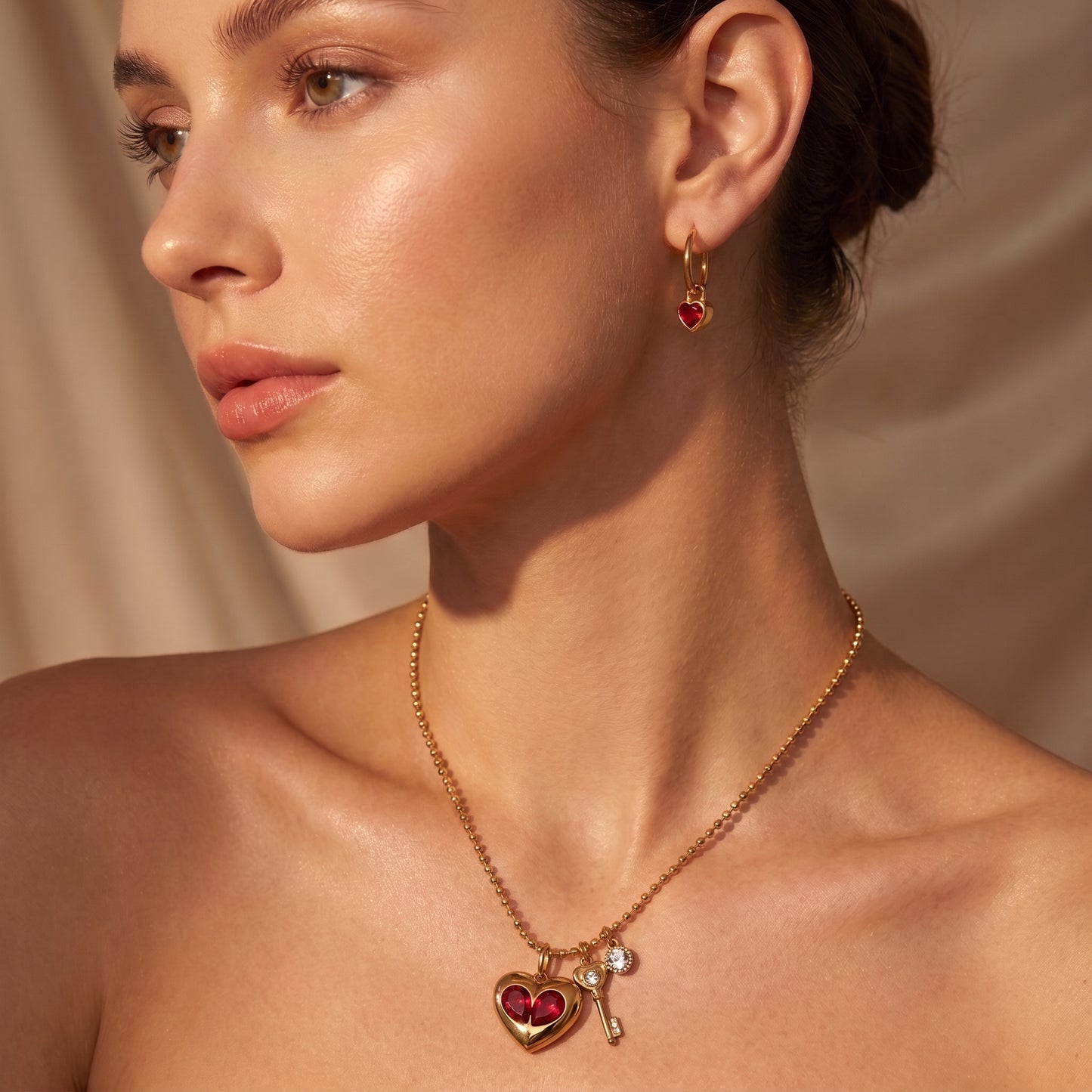 A close-up of a woman wearing a Sleek gold Amour Necklace featuring red crystal heart inserts, a golden key charm, and a clear crystal and matching drop earrings, set against a soft beige fabric background.