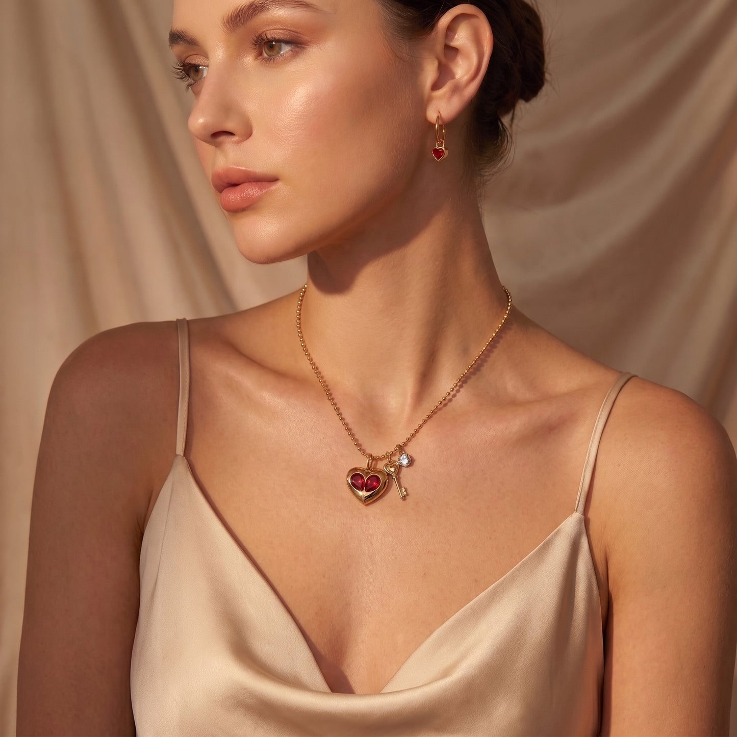 A woman wearing a Sleek gold Amour Necklace featuring red crystal heart inserts, a golden key charm, and a clear crystal and a matching drop earring, styled with a satin camisole against a draped beige fabric background.