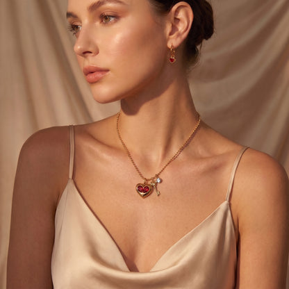 A woman wearing a Sleek gold Amour Necklace featuring red crystal heart inserts, a golden key charm, and a clear crystal and a matching drop earring, styled with a satin camisole against a draped beige fabric background.