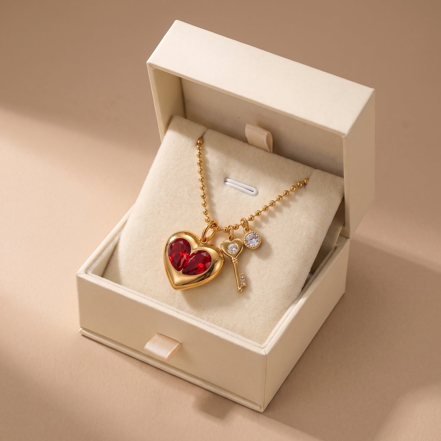 Sleek gold Amour Necklace featuring red crystal heart inserts, a golden key charm, and a clear crystal, displayed inside an open cream-colored jewelry box on a beige background.