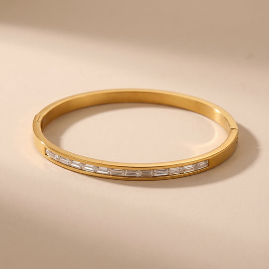 High quality gold Aria Bracelet featuring channel-set clear baguette crystals resting on travertine stone, highlighting its tarnish-free and skin friendly finish, displayed on a warm beige background.