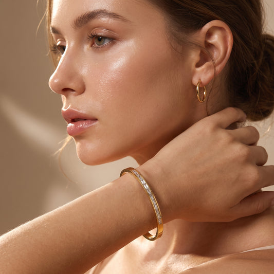 A close-up lifestyle shot of a woman wearing a slim High quality gold Aria Bracelet featuring channel-set clear baguette crystals resting on travertine stone, highlighting its tarnish-free and skin friendly finish, set against a warm beige background.