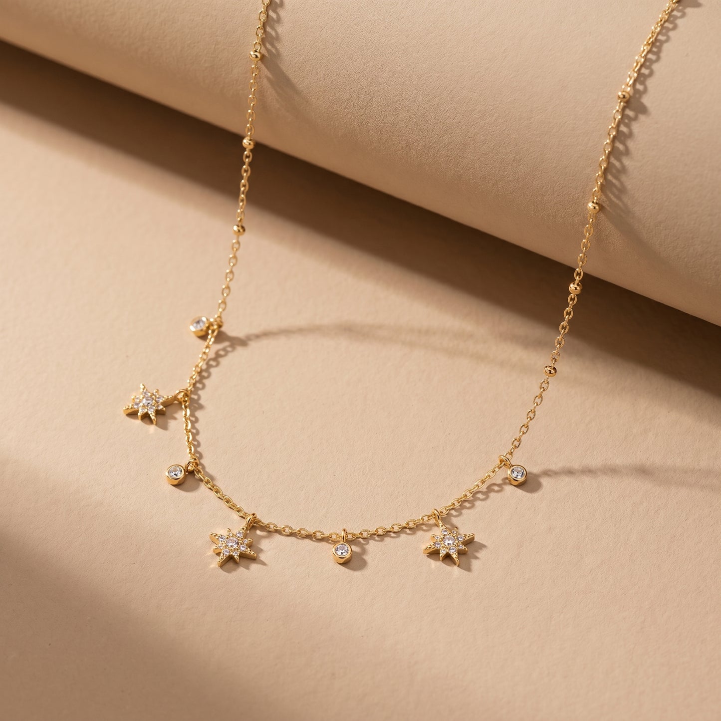 Dainty gold chain necklace featuring alternating dangling starburst charms and clear crystal drops, highlighting its tarnish-free and skin friendly finish. on a beige background.