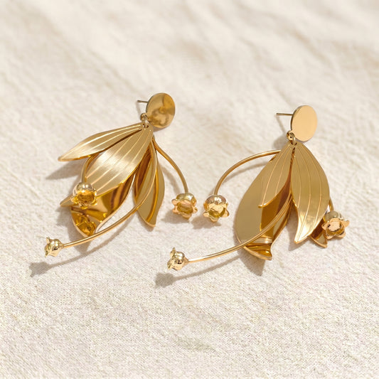 High quality waterproof gold plated statement earrings featuring a modern botanical design with sweeping leaves and cascading lily flowers, laid flat on a pristine background