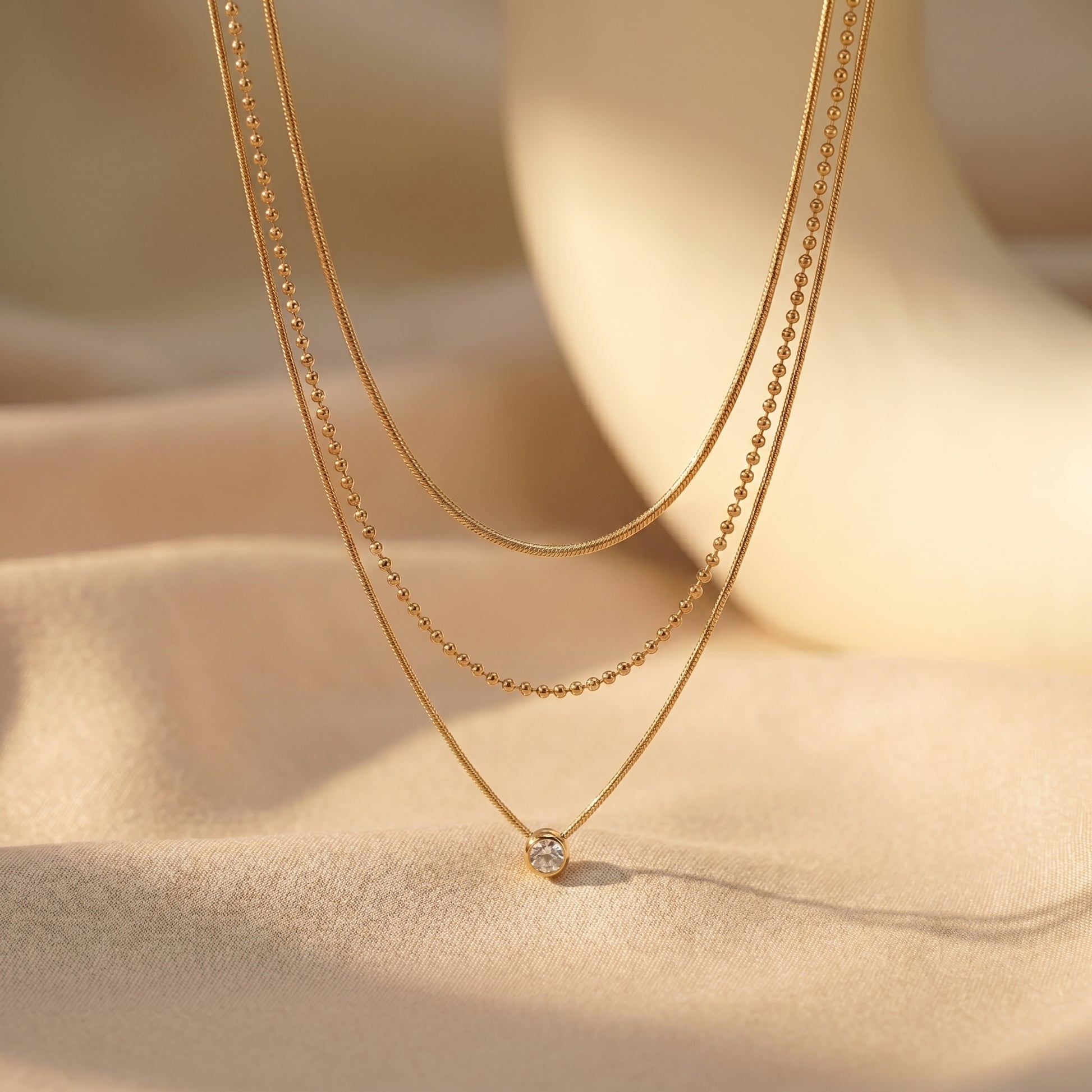 High quality waterproof gold plated triple layer necklace featuring delicate snake chains, a beaded chain, and a solitaire crystal pendant on a beige background