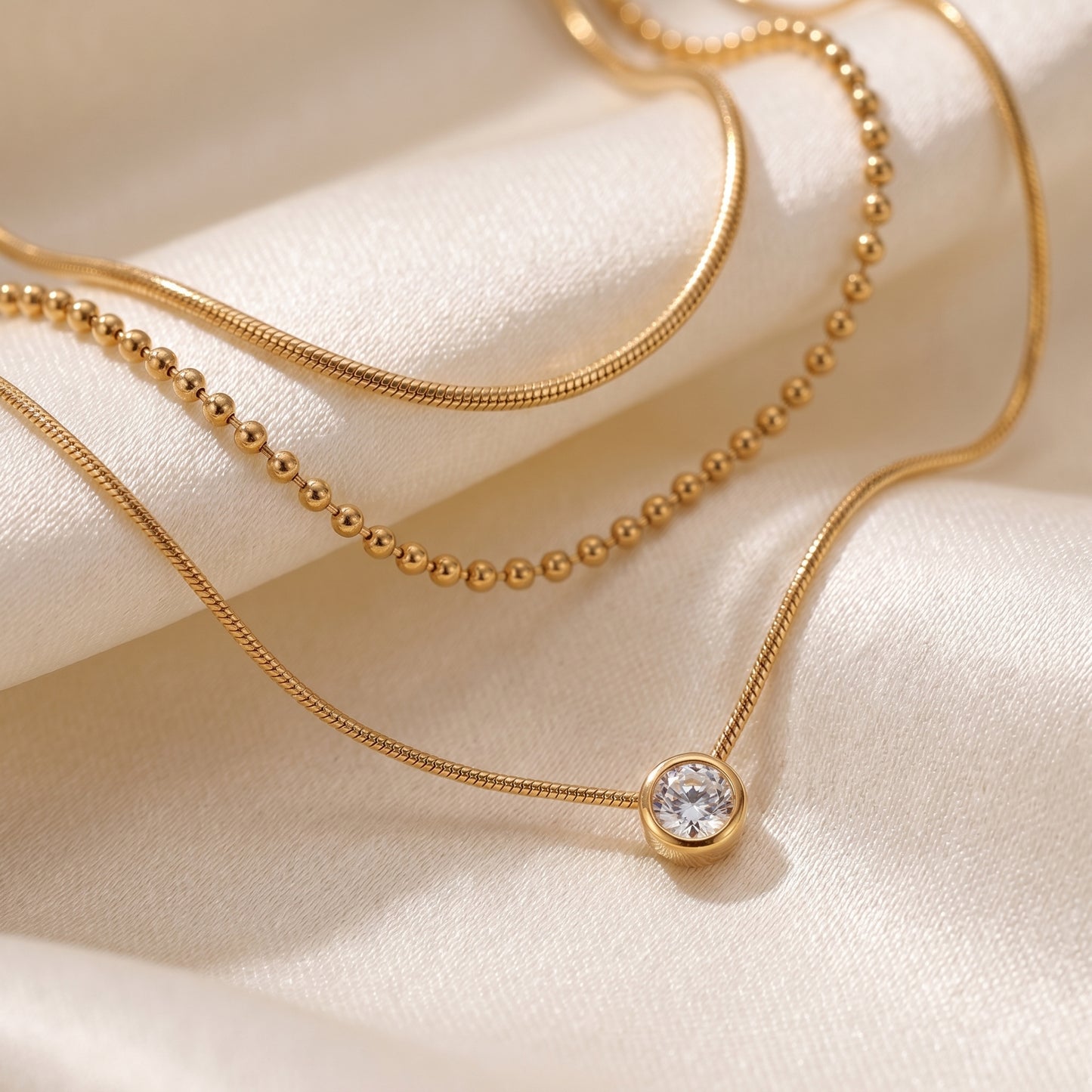 High quality waterproof gold plated triple layer necklace featuring delicate snake chains, a beaded chain, and a solitaire crystal pendant on a beige fabric background