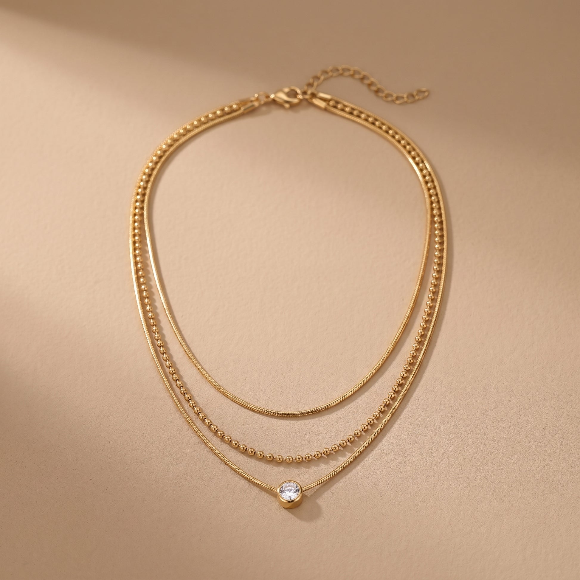 High quality waterproof gold plated triple layer necklace featuring delicate snake chains, a beaded chain, and a solitaire crystal pendant on a beige background.