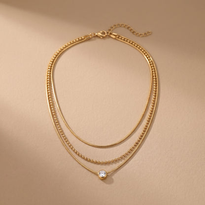 High quality waterproof gold plated triple layer necklace featuring delicate snake chains, a beaded chain, and a solitaire crystal pendant on a beige background.