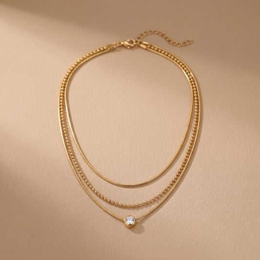 High quality waterproof gold plated triple layer necklace featuring delicate snake chains, a beaded chain, and a solitaire crystal pendant on a beige background.
