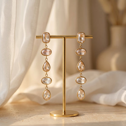 Gold earrings with clear gemstones on a gold stand against a soft, neutral background