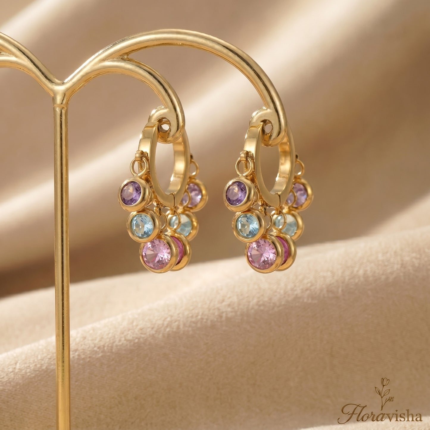Gold hoop earrings with colorful gemstones on a beige background, featuring the brand 'Floravisha'.