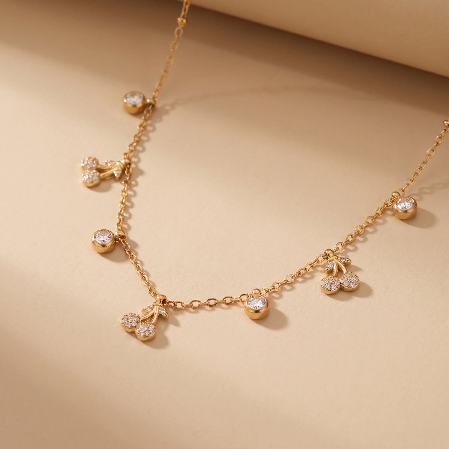 Elegant dainty gold necklace featuring alternating bezel-set crystals and micro-pave cherry charms on a beige background.