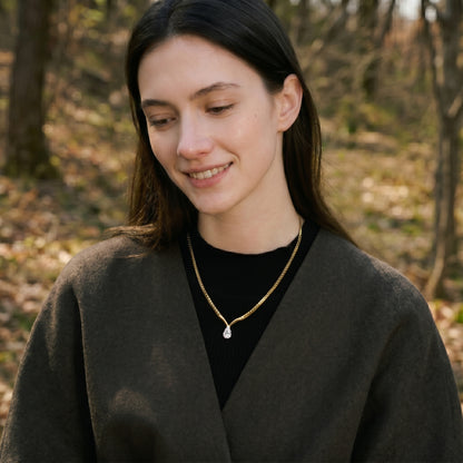 Woman wearing a dark coat and black shirt with a gold necklace in an outdoor setting.