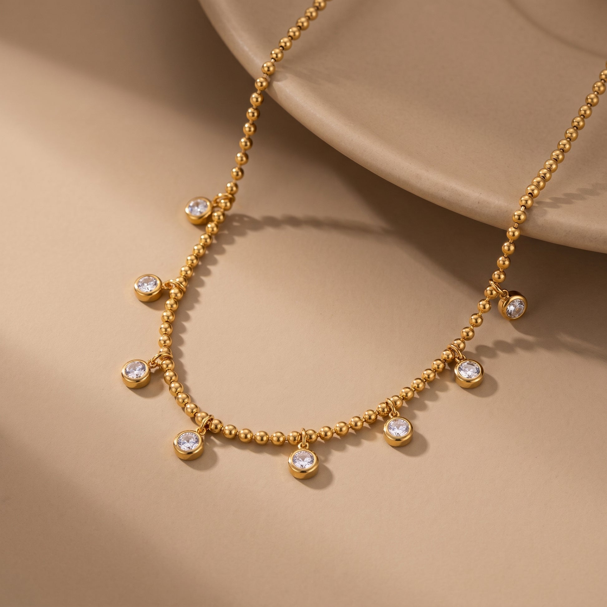 Elegant, daintier gold-beaded satellite chain necklace featuring alternating dangling bezel-set clear crystals. highlighting its tarnish-free and skin friendly finish on a beige background.