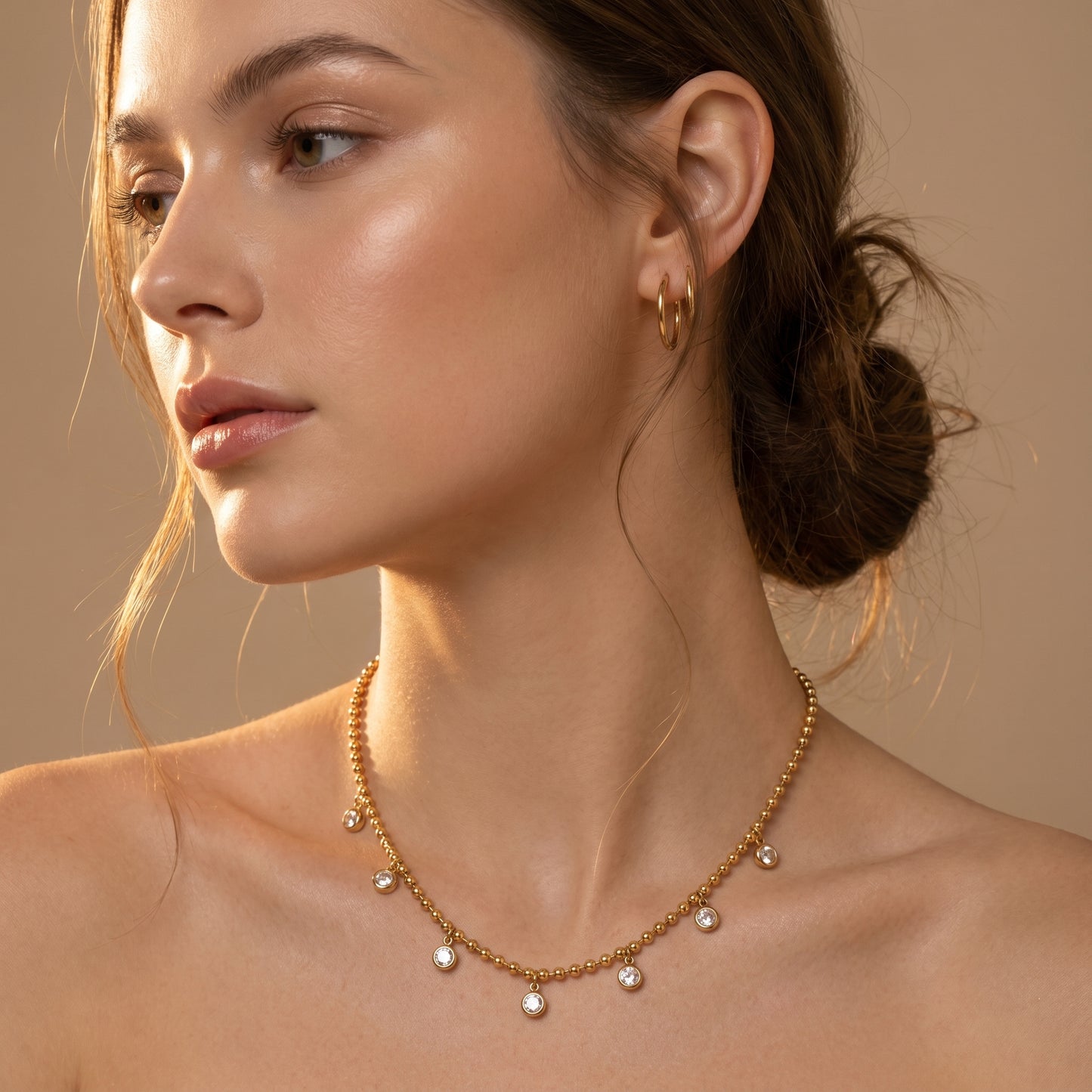 Woman wearing Elegant, daintier gold-beaded satellite chain necklace featuring alternating dangling bezel-set clear crystals on a clean white background, highlighting its tarnish-free and skin friendly finish against a beige background.
