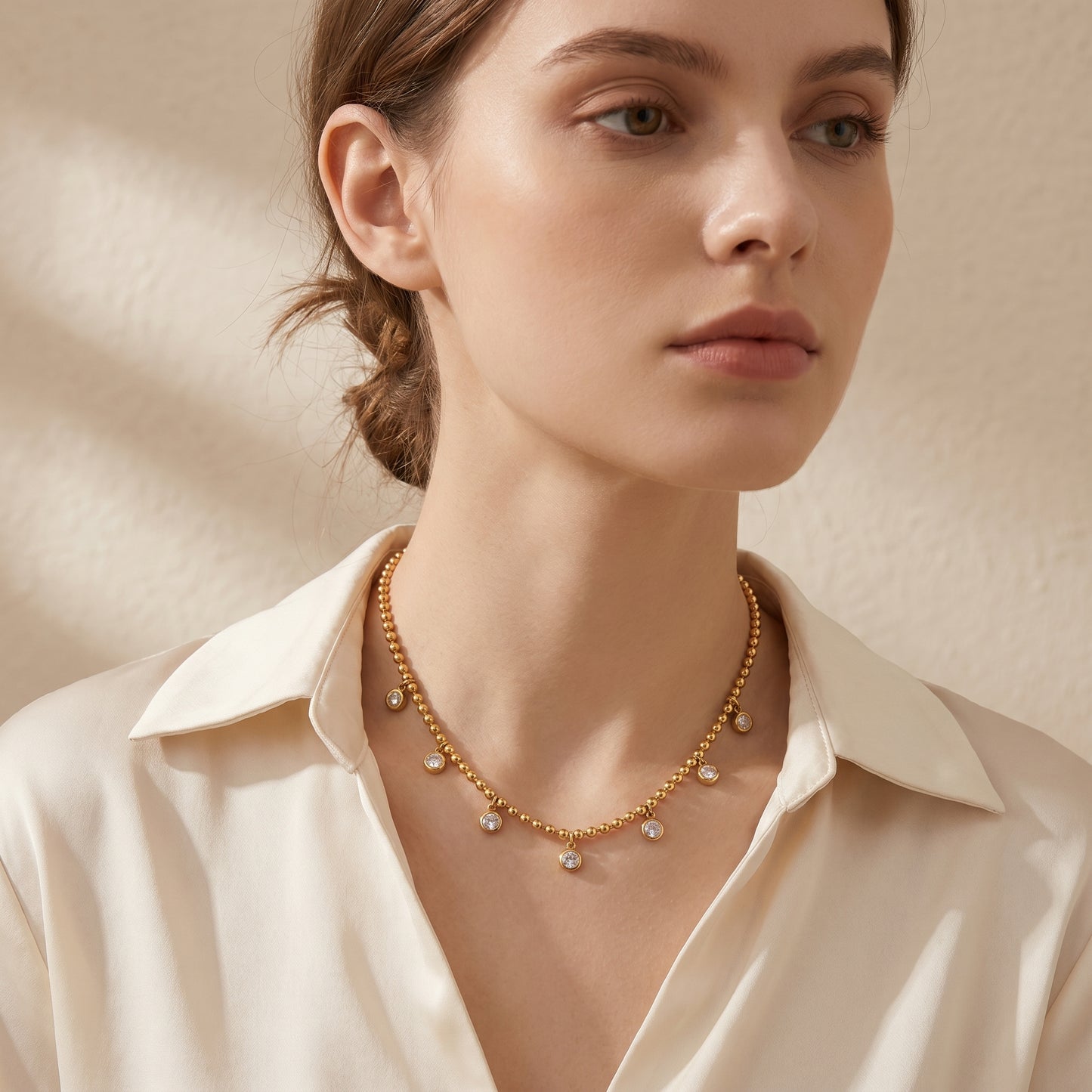 Woman wearing a Elegant, daintier gold-beaded satellite chain necklace featuring alternating dangling bezel-set clear crystals on a clean white background, highlighting its tarnish-free and skin friendly finish with a beige background