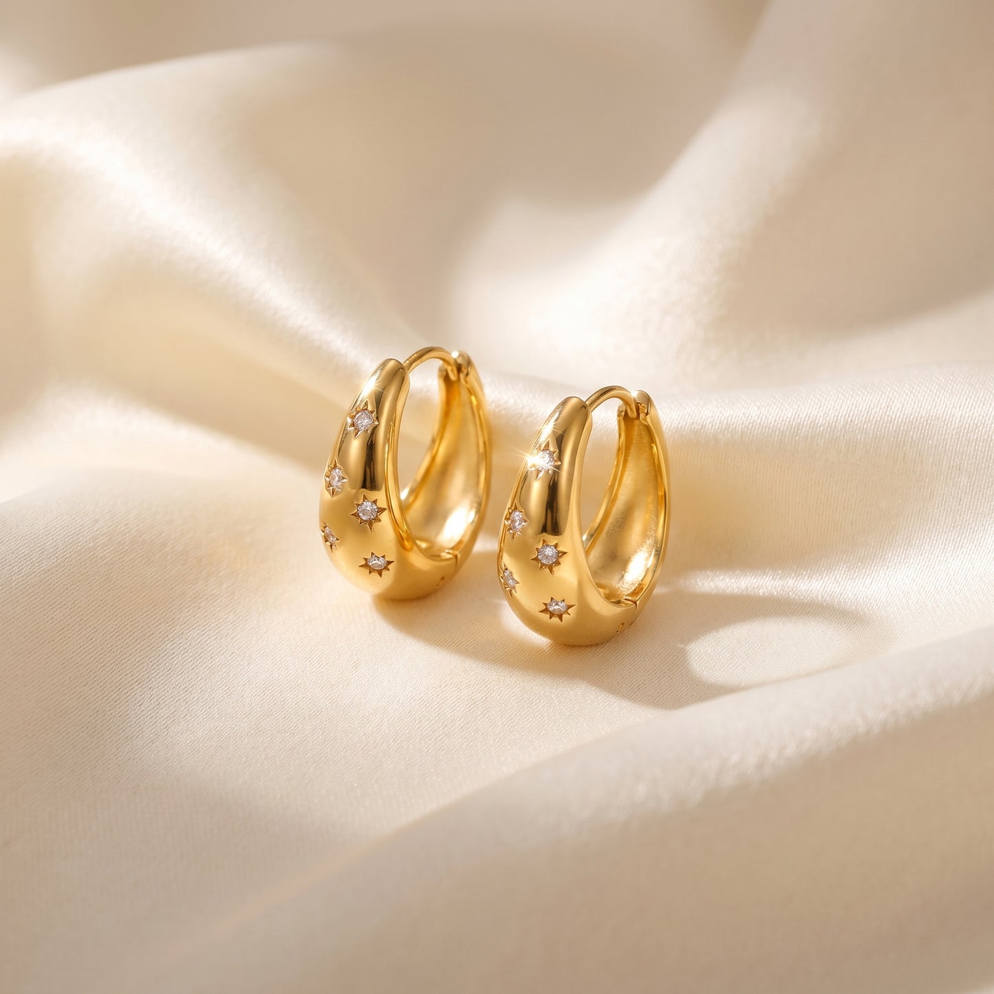 High quality waterproof gold plated chunky dome hoop earrings with engraved starburst crystals, shown standing with a soft reflection on a beige fabric background
