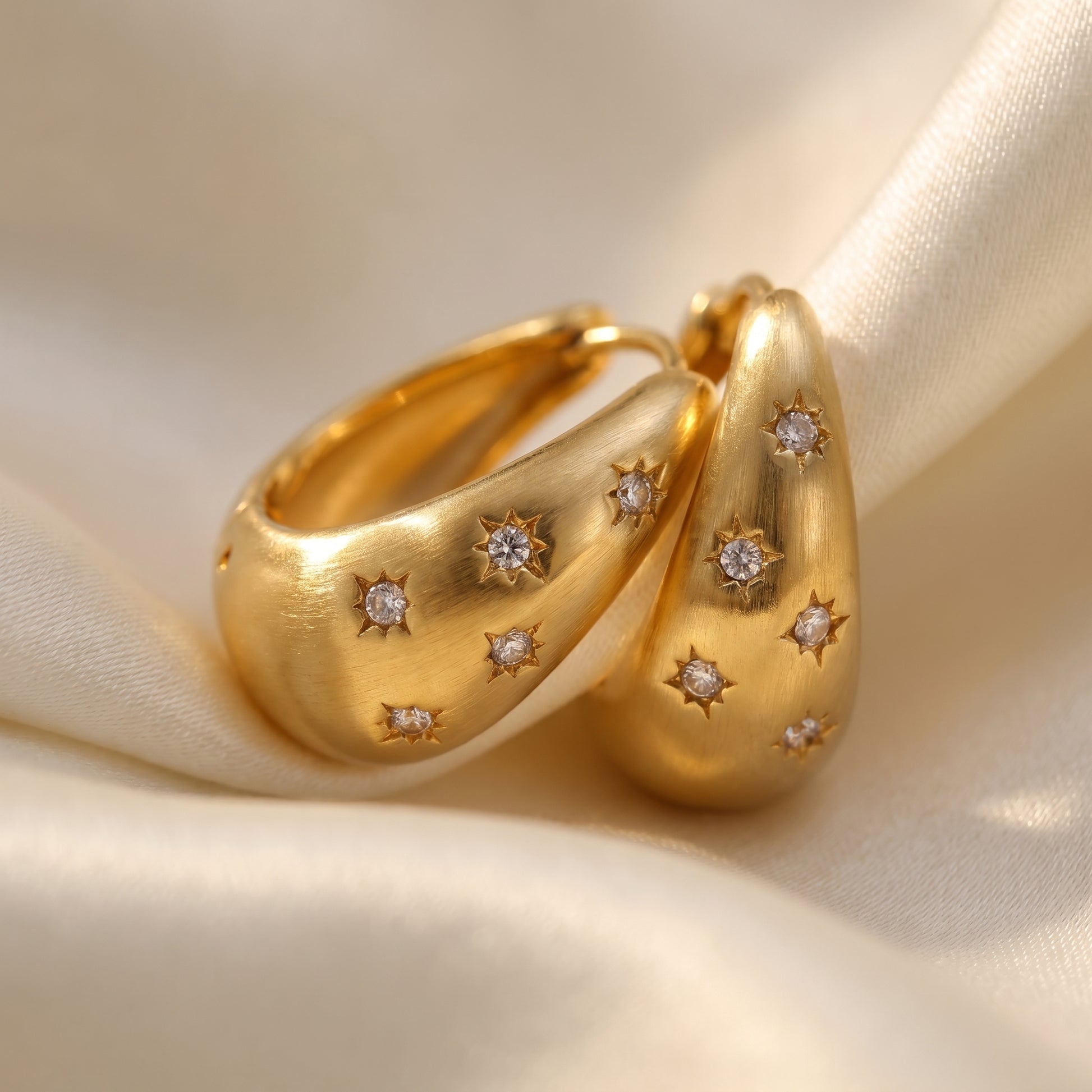 High quality waterproof gold plated chunky dome hoop earrings with engraved starburst crystals, shown standing with a soft reflection on a beige fabric background.