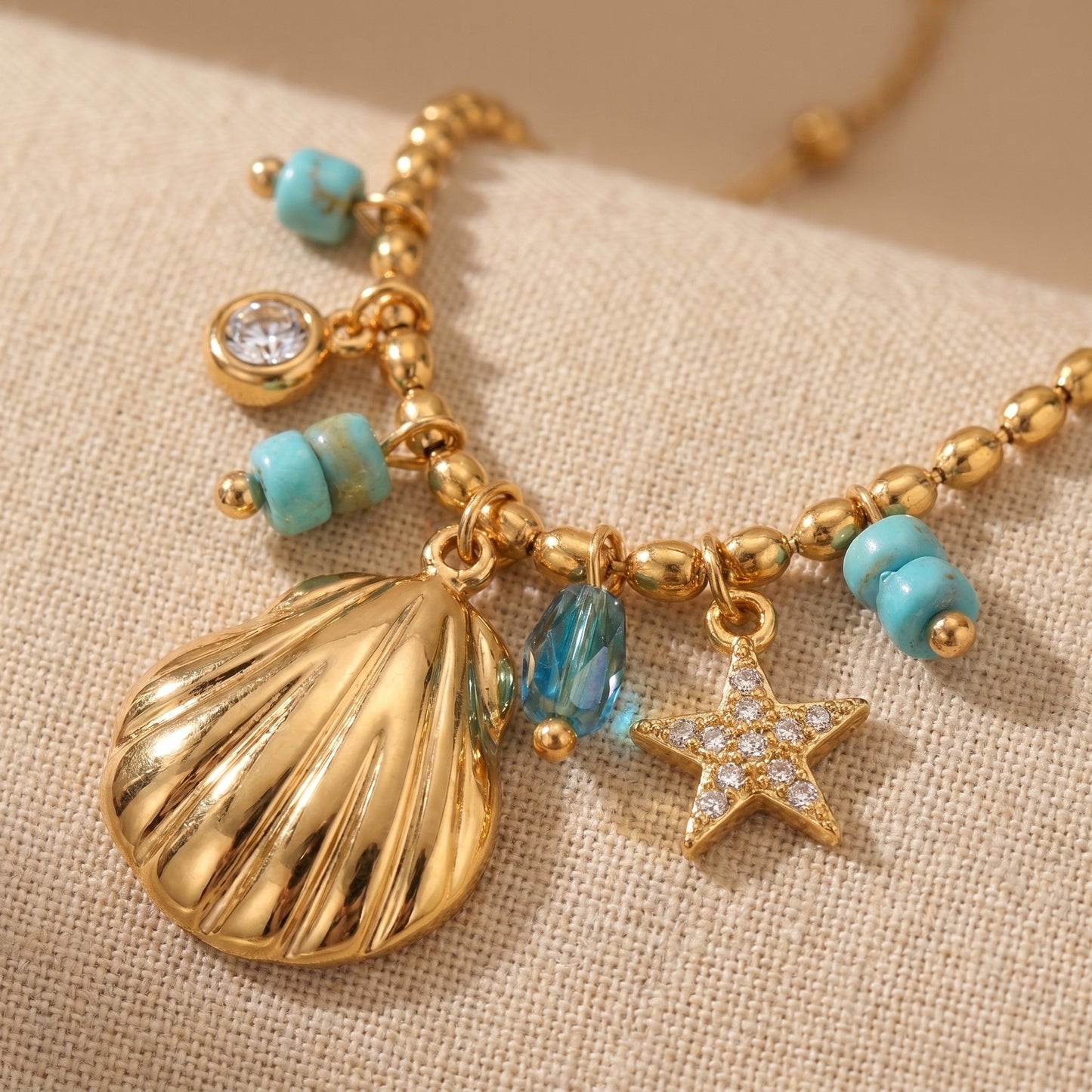 Gold necklace with shell, starfish, and turquoise bead charms on a beige fabric background