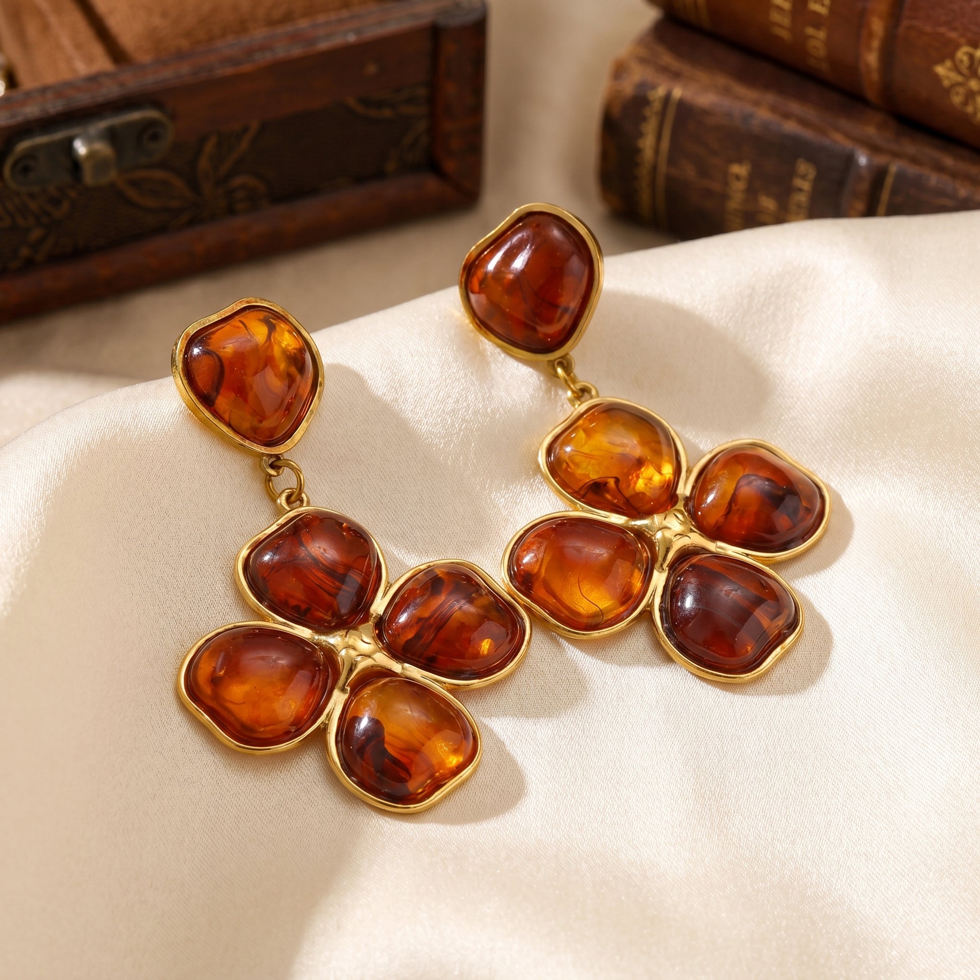 Close-up of luxury amber floral drop earrings resting on a warm beige matte surface, highlighting the tarnish-free gold plating and rich tortoiseshell resin details.