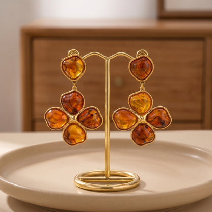 Close-up of luxury amber floral drop earrings resting on a warm beige matte surface, highlighting the tarnish-free gold plating and rich tortoiseshell resin details.