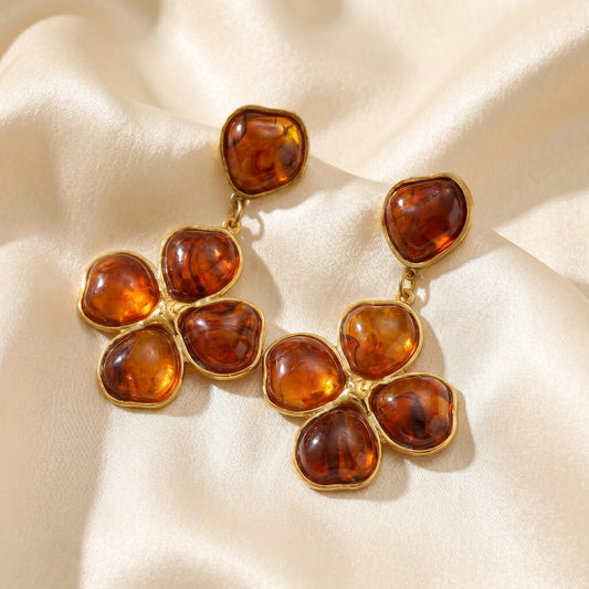Close-up of luxury amber floral drop earrings resting on a warm beige matte surface, highlighting the tarnish-free gold plating and rich tortoiseshell resin details.
