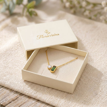 Gold necklace with a green pendant in an open jewelry box labeled 'Floravisha' on a textured surface.