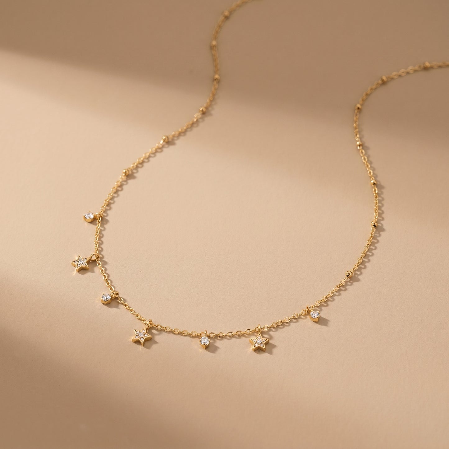 Dainty gold beaded satellite chain necklace featuring alternating dangling mini starburst charms and clear crystal drops on a clean white background, highlighting its tarnish-free and skin friendly finish on a beige background