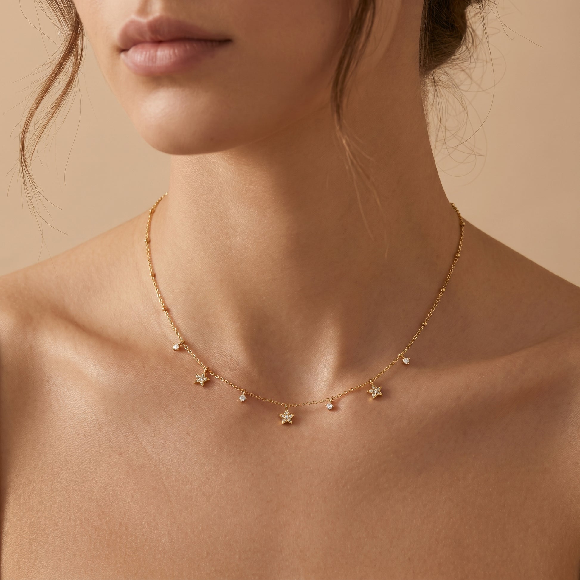 Close-up of a person wearing a Dainty gold beaded satellite chain necklace featuring alternating dangling mini starburst charms and clear crystal drops, highlighting its tarnish-free and skin friendly finish on a beige background.