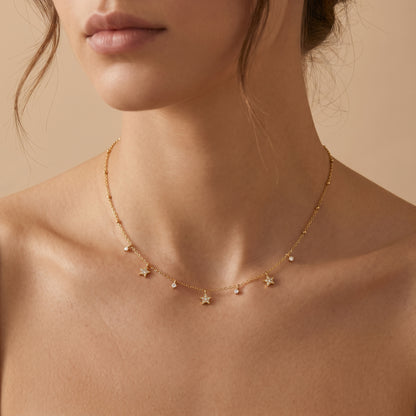 Close-up of a person wearing a Dainty gold beaded satellite chain necklace featuring alternating dangling mini starburst charms and clear crystal drops, highlighting its tarnish-free and skin friendly finish on a beige background.