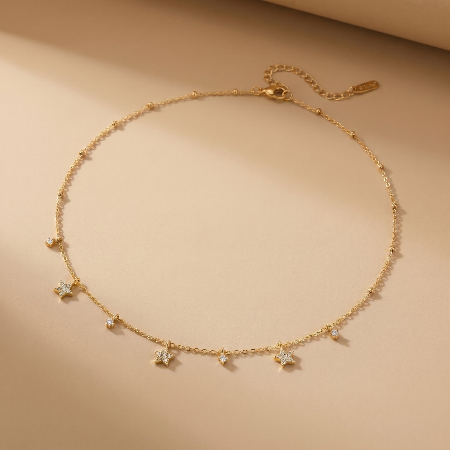 Dainty gold beaded satellite chain necklace featuring alternating dangling mini starburst charms and clear crystal drops. highlighting its tarnish-free and skin friendly finish on a beige background