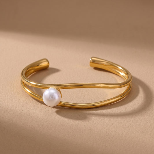 The tarnish-free Oceane Wave Pearl Cuff Bangle by Floravisha, displaying two fluid asymmetrical gold bands wrapping a lustrous white pearl on a clean background.