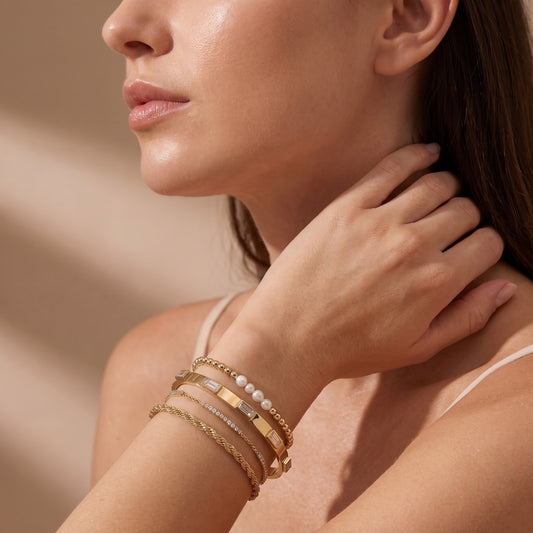 Modern gold wrist stack on a woman's arm showcasing the waterproof Verona bezel crystal bracelet layered with a pearl bead chain, diamond tennis chain, and twisted rope cuff.