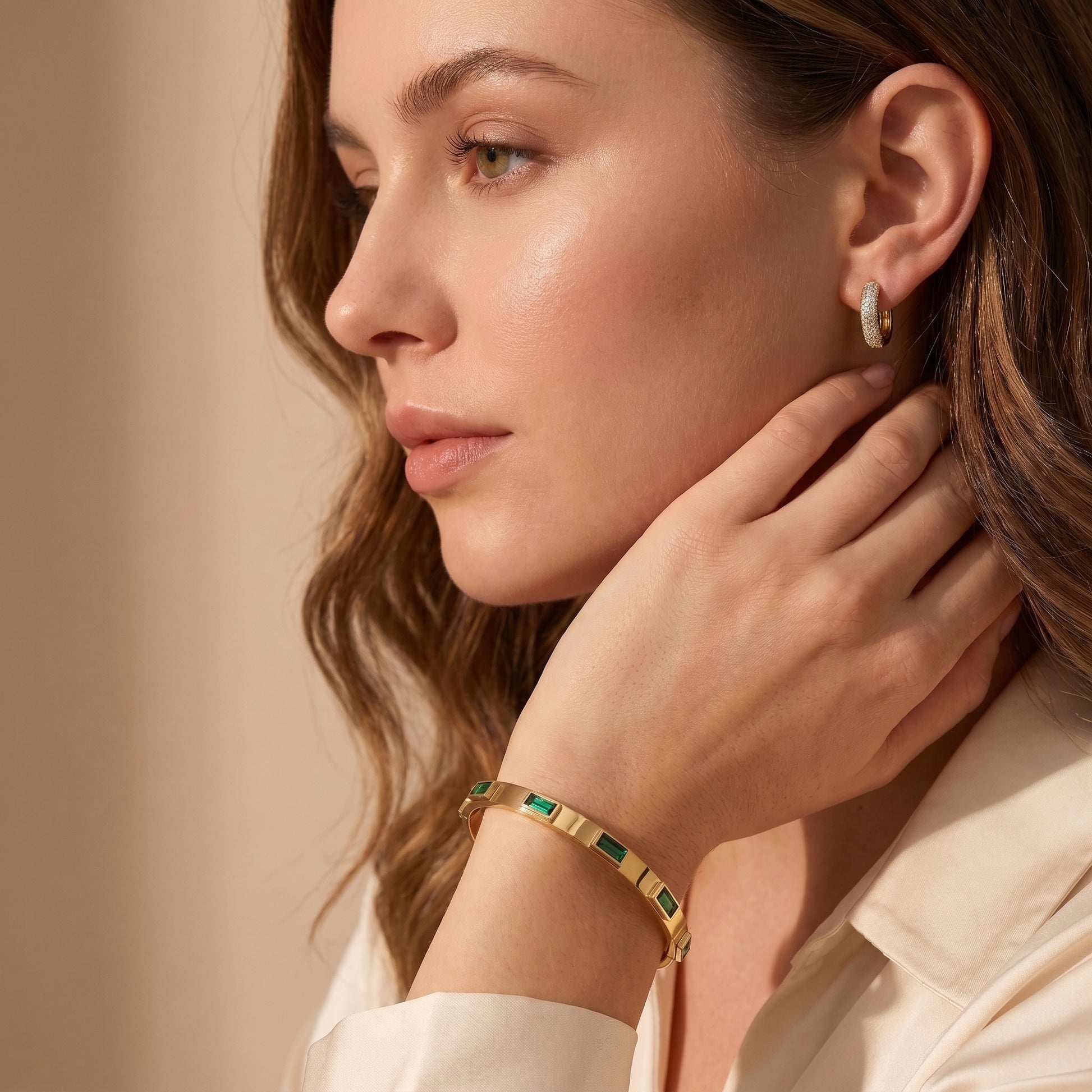 Close-up side profile of a woman wearing the skin friendly Verona gold bezel bracelet and crystal huggie studs with a cream silk blouse.