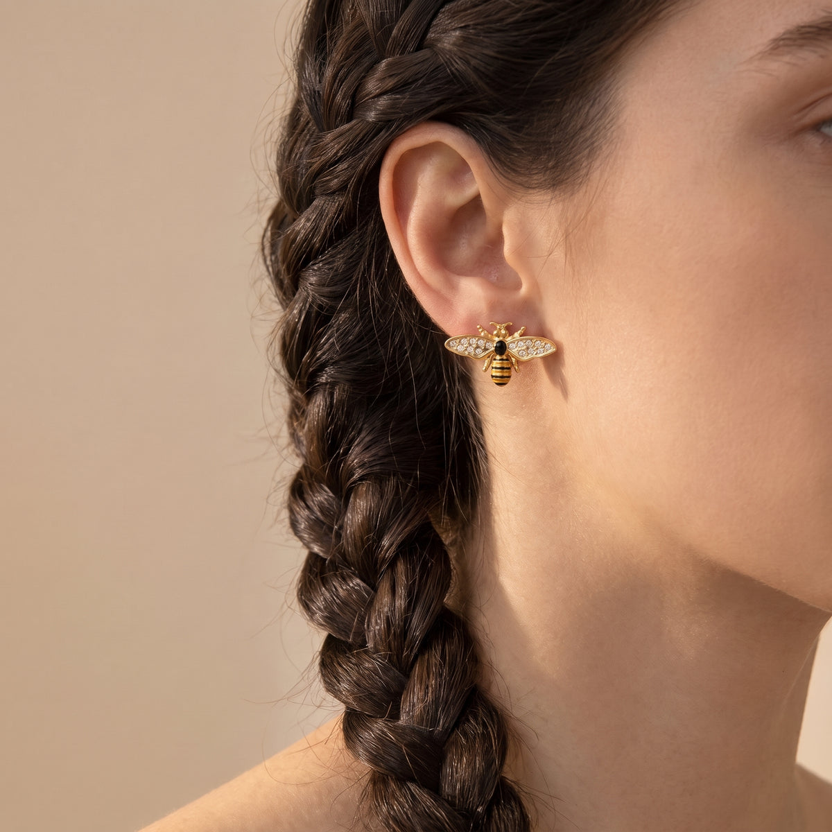 Macro profile view of a woman with braided hair wearing the hypoallergenic Abeille gold honeybee crystal stud earring resting flush on the earlobe.