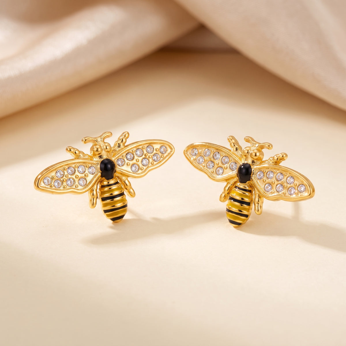 High quality Abeille gold pavé honeybee stud earrings by Floravisha showing clear cubic zirconia wings and striped enamel bodies on a warm neutral surface.