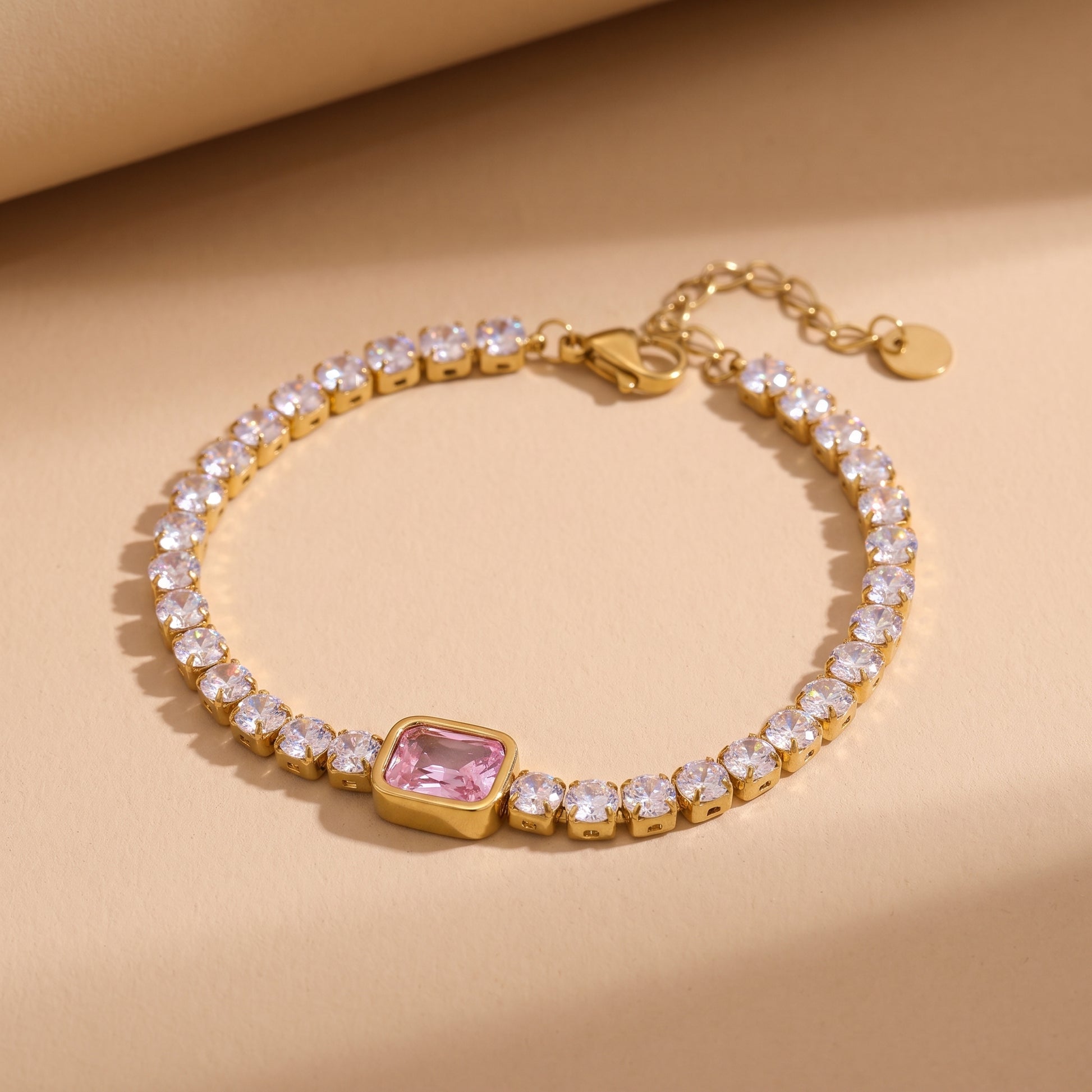 High quality Amara Gold Tennis Bracelet by Floravisha featuring clear round crystals and a center pink emerald-cut gemstone on a warm beige studio background.